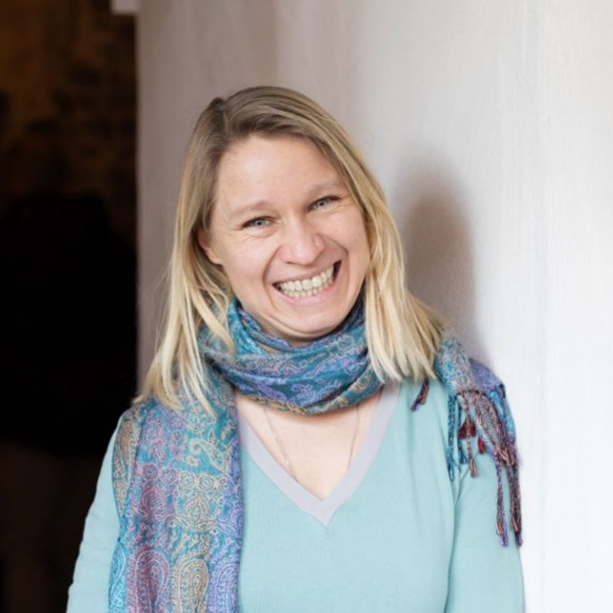 Lächelnde Frau mit blonden Haaren und buntem Schal vor einer hellen Wand.